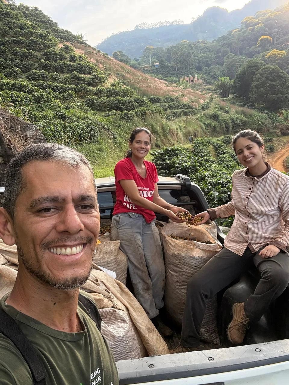 Cassiano em primeiro plano na caçamba da caminhonete, com Adélia e Maria Clara sentadas sobre os sacos de café recém-colhido, mostrando as cerejas nas mãos, com as lavouras e a serra ao fundo